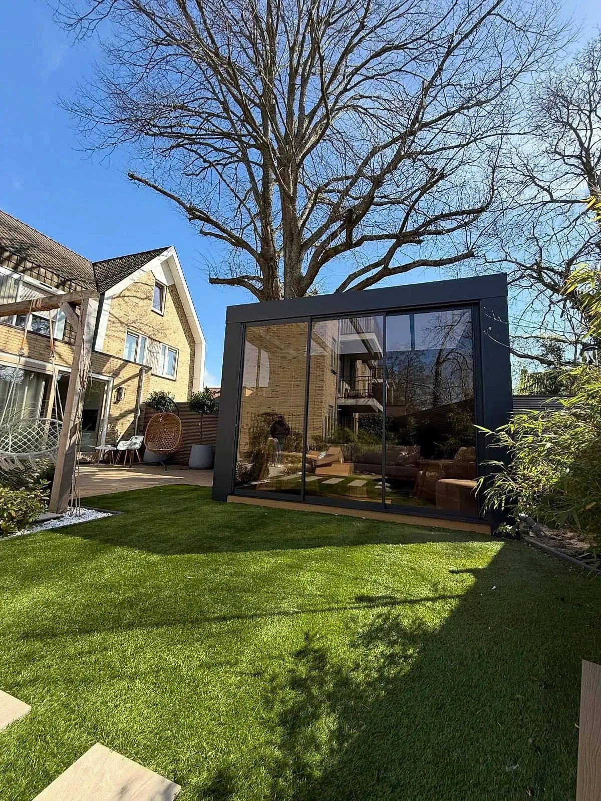 Antraciet cube met drie glazen schuifpanelen achter in een tuin met gras en een grote boom