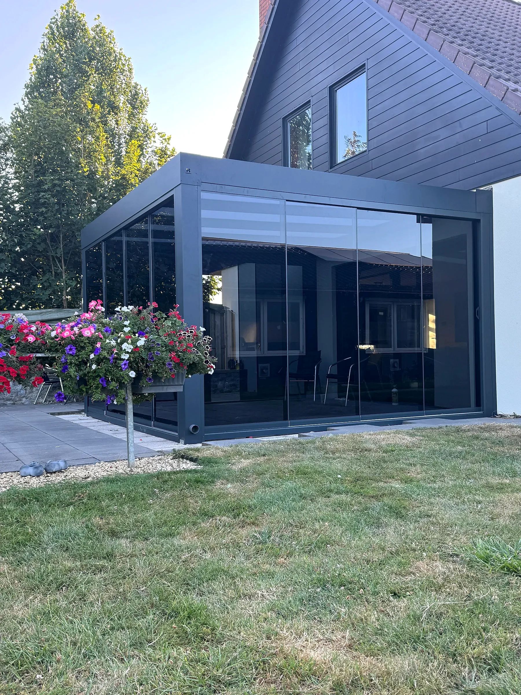 Antraciet cube met glazen schuifwanden aan een woning, met bloeiende hanging baskets ervoor