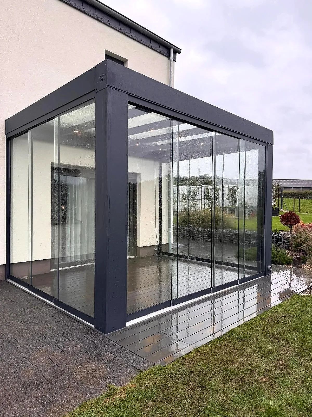 Antraciet cube met heldere glazen schuifwanden op een terras naast een lichte woning