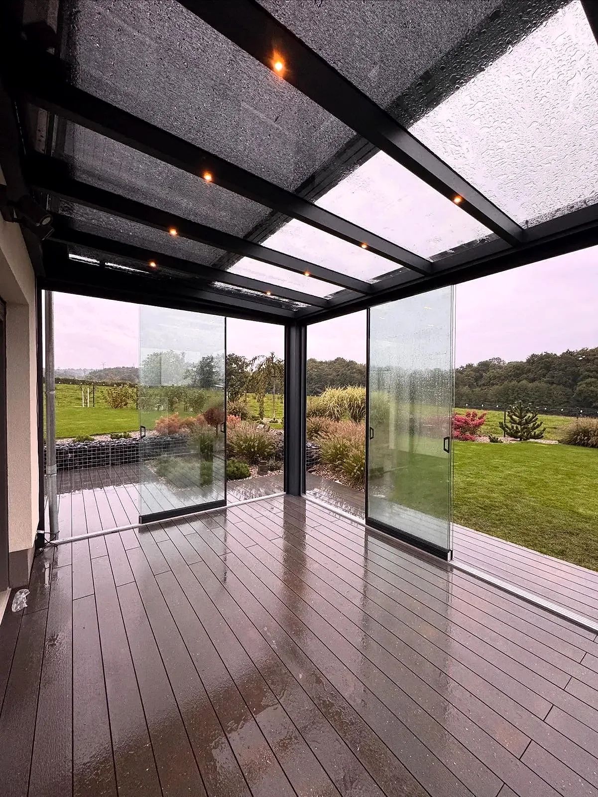 Zicht vanuit een cube met lichtspots in het dak door heldere glazen schuifwanden naar de tuin