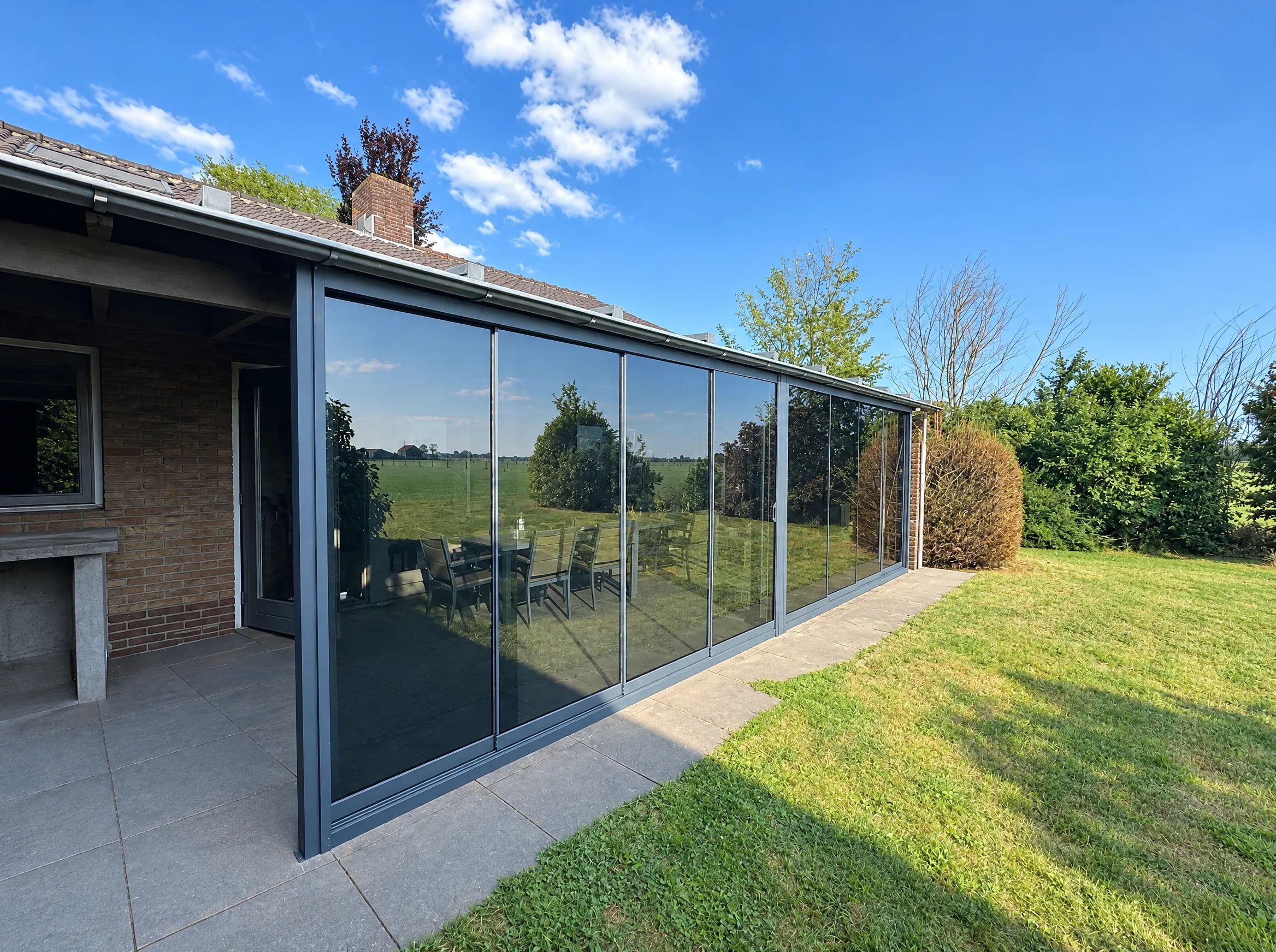 Lange rij getinte glazen schuifwanden onder de dakgoot van een bungalow, met gazon ervoor en weilanden die in het glas spiegelen