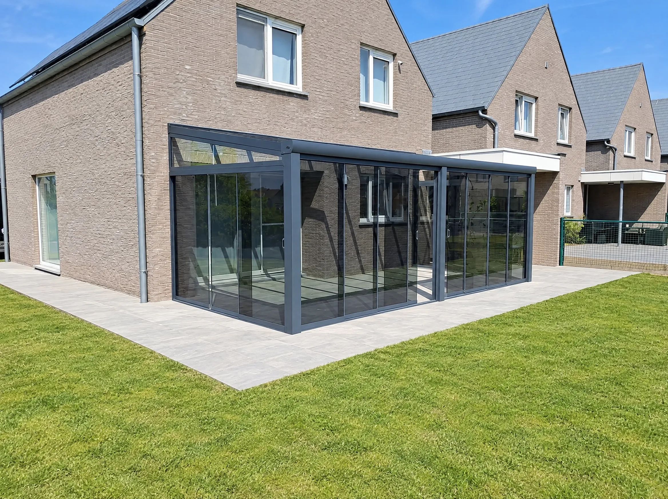 Hoekveranda met getinte glazen schuifwanden aan de voor- en zijkant, aan een bakstenen woning met gazon
