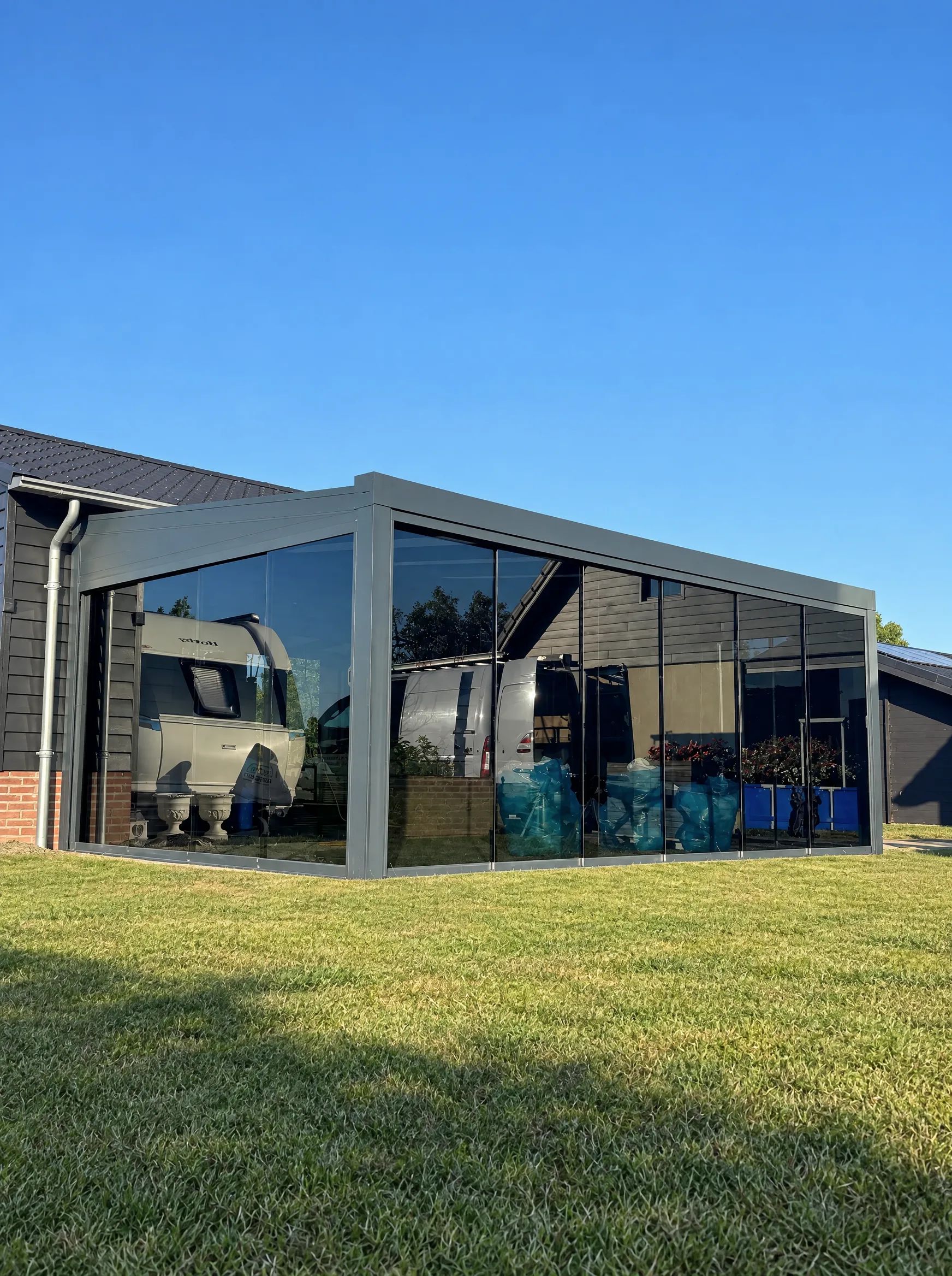 Grijze veranda met getinte glazen schuifwanden aan twee zijden, naast een woning met donkere gevelbekleding