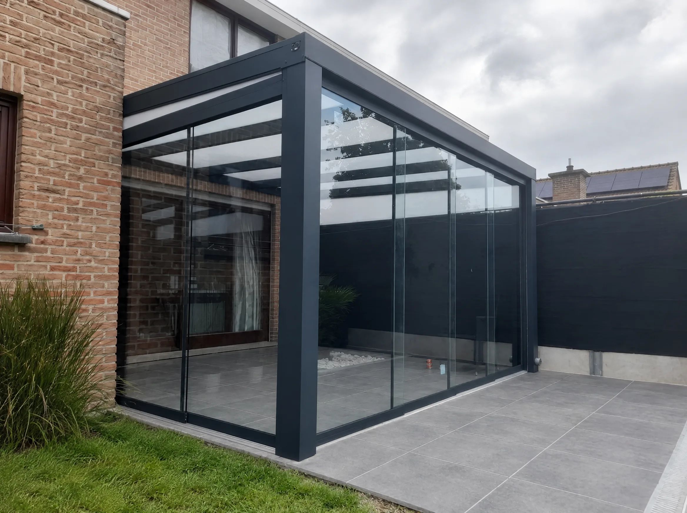Antraciet veranda op een tegelterras, aan twee zijden afgesloten met heldere glazen schuifwanden